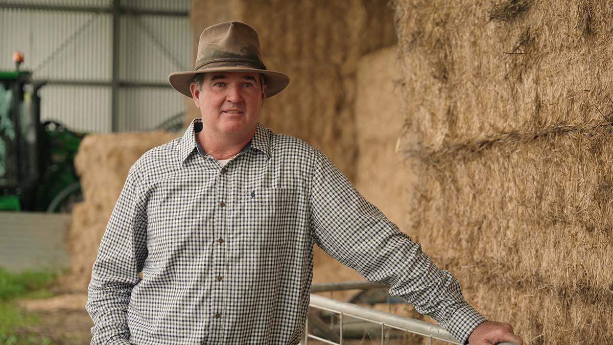 Brad Nicholson wearing a wide-brimmed hat and checked shirt, leaning on a metal gate next to stacked hay bales